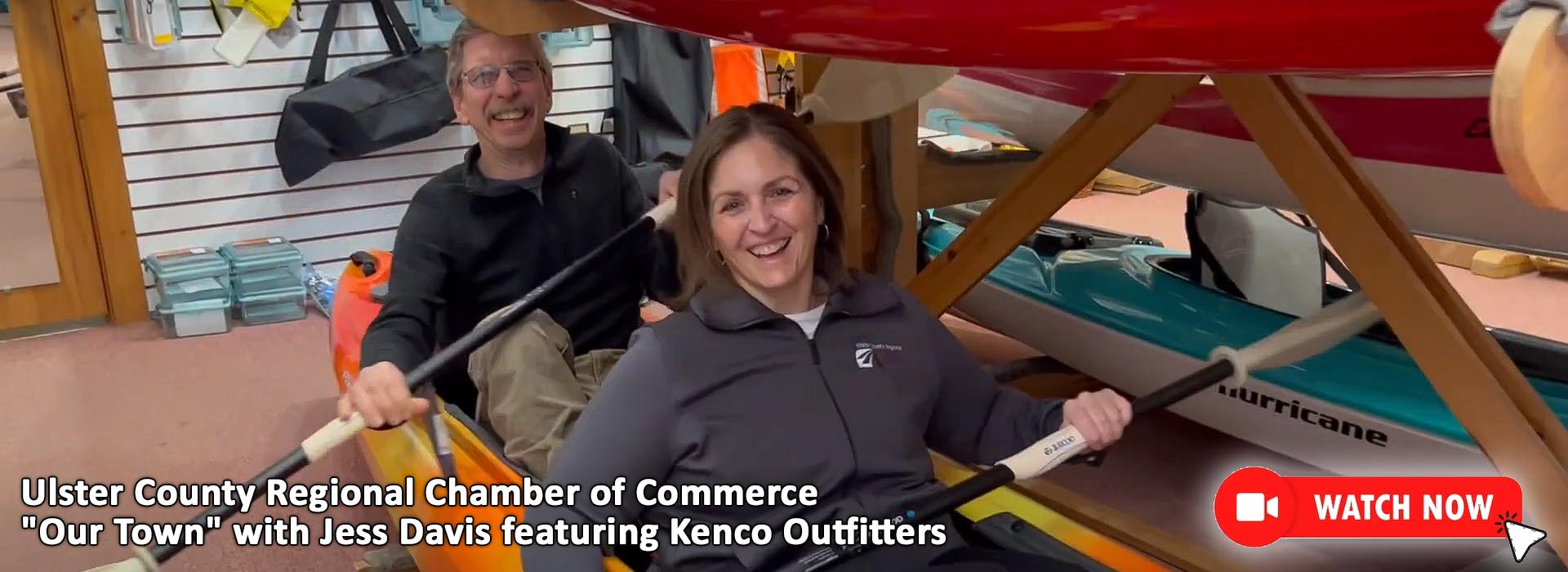 UCRCC Our Townm Featuring Kenco Outfitters. Store owner Bill Kennedy and Ulster County Regional Chamber of Commerce Director of Membership Jess Davis are seated in an orange, red and yellow swirled kayak holding kayaking paddles and pretending to kayak on our showroom floor. Bill is wearing glasses, a black halfzip fleece pullover and olive cargo pants. Jess is wearing a navy blue Ulster Chamber of Commerce fleece jacket and dark pants. They are both smiling. In the lower right corner is a video icon and watch now button. Clicking on this image will open a page to watch the Our Town episode on Kenco Outfitters